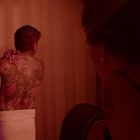 Keine Texte sichtbar. Ein tätowierter Mann mit Handtuch steht mit dem Rücken zur Kamera; eine Frau schaut zu. Warmes Licht.