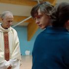 Ein älterer Mann im Messgewand mit rotem und goldenen Muster steht, während ein Jugendlicher und eine Frau im Hintergrund unscharf sind.