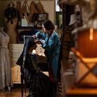 Eine Frau arbeitet konzentriert an einer sitzenden Person in historischer Kleidung, umgeben von Schneiderbedarf und Kostümen.