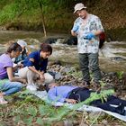 Vier Personen knien an einem Fluss neben einer Person, die auf dem Boden liegt. Eine Person trägt ein "Polizei"-Band.