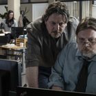 Zwei Männer arbeiten konzentriert am Computer in einem Büro; einer sitzt, der andere steht daneben und schaut mit.