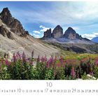 Text: Italien, Südtirol, Drei Zinnen. 
Berglandschaft mit massiven Felsformationen und lila Blumen im Vordergrund.