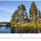 "Häuser am See". Ein Kalenderbild zeigt rote Holzhäuser am Ufer eines Sees, umgeben von hohen Kiefern und klarem blauem Himmel.