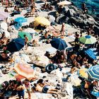 Viele Menschen sonnen sich auf Felsen neben dem Meer. Bunte Sonnenschirme bieten Schatten.