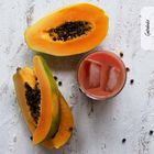 Text "Getränke" oben rechts. Zwei halbe Papayas und ein Glas roter Saft mit Eiswürfeln auf weißer Oberfläche.