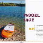 "Paddel Ahoi! ... auf der Kollerinsel am Rhein #41" Ein See mit einem orange-weißen Paddelboard am sandigen Ufer.