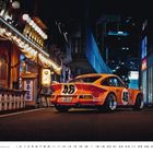 Ein orangefarbenes Rennfahrzeug mit der Nummer 46 steht nachts auf einer Straße, umgeben von beleuchteten Gebäuden.