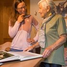 Zwei Frauen stehen an einem Tisch mit einem geöffneten Buch, eine junge und eine ältere Frau, beide lächeln einander an.