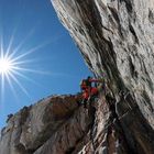 Ein Kletterer in roter Ausrüstung erklimmt eine steile Felswand unter klarem, blauem Himmel und strahlender Sonne.