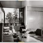 Ein älterer Mann sitzt auf einem Sofa in einem modernen Raum mit großen Fenstern, Büchern und Pflanzen.