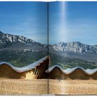 Eine wellenförmige Architektur vor einer Gebirgskulisse mit blauem Himmel, abgebildet in einem geöffneten Buch.
