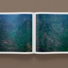 Ein aufgeschlagenes Buch zeigt ein lebhaftes Bergtal mit grünen Wäldern und steilen Felswänden.