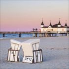 "9", "OM 42", "42" auf Strandkörben am Sandstrand, weiße Seebrücke mit Türmen im Hintergrund, rosa-blauer Himmel.