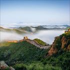 Eine lange Mauer schlängelt sich über grüne Hügel im Nebel, umgeben von üppiger Vegetation und blauem Himmel.