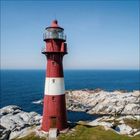 Leider sind keine Texte zu sehen. Ein roter und weißer Leuchtturm steht auf felsiger Küste vor blauem Meer unter klarem Himmel.