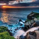 Wasserfall an einer felsigen Küste bei Sonnenuntergang, Meer unter bewölktem Himmel, Licht spiegelt auf Wasser.