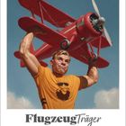 "FlugzeugTräger" steht über einem muskulösen Mann, der ein rotes Doppeldecker-Flugzeug hebt. "Juli 2027" ist der Monat.