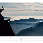 Kalenderseite für Oktober mit zwei Steinböcken auf einer Bergklippe, Wolkenmeer und Bergsilhouetten im Hintergrund.