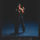 Oktober  |  October. Zwei Tänzer stehen nahe beieinander, einer hält eine Kerze, umgeben von Dunkelheit.