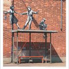 (Text: "Clear Channel") Obdach mit Graffiti von tanzenden Figuren auf roter Backsteinmauer. Darunter ein leerer roter Sitz.