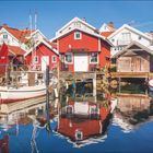 Kalender für Juli und August, Text über Falunrote Häuschen. Bild zeigt rote Holzhäuser und Boote an einem sonnigen Hafen.