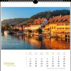 Oktoberkalender mit Wochenübersicht. Bild: Malerische Häuserreihe am Wasser bei Sonnenuntergang, umgeben von Waldhügeln.