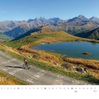 Oktober. Ein Radfahrer auf einer Bergstraße, umgeben von hohen, felsigen Bergen und einem ruhigen See.