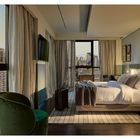 Ein elegantes Zimmer mit großem Bett, Fenstern mit Vorhängen, Fernseher, kleinem Tisch, Sessel und einer Reisetasche.