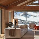 Keine Texte. Moderner Raum mit Holzbalkendecke, großen Fenstern, beigem Sofa, Sessel, Tisch und Blick auf verschneite Berge.