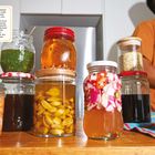 Texte über zwei Reihen von eingelegten Speisen, daneben diverse bunte Gläser mit Inhalt. Links blauer Stoff.