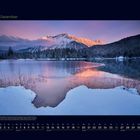 12 Dezember. Berge im Sonnenuntergang spiegeln sich in einem teils gefrorenen See, umgeben von verschneiten Wäldern.