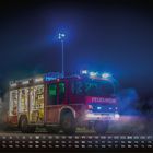 Text: "FEUERWEHR", Kalenderdaten unten. Ein Feuerwehrauto mit offenen Fächern, beleuchtet in der Nacht, umgeben von Rauch.