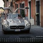 Ein Silberner Mercedes-Benz 300 SL mit Flügeltüren fährt durch eine enge Straße; Menschen und historische Gebäude im Hintergrund.