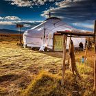 Eine weiße Jurte steht in einer grasbewachsenen Landschaft unter einem bewölkten Himmel. Links zieht eine Person Vieh.