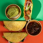 Zwei gefüllte Tacos auf rotem Hintergrund, eines stehend auf grünem Hintergrund. Schüsseln mit Guacamole und Salsa.