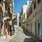 Steile, gepflasterte Straße zwischen hellen Gebäuden mit Balkonen in mediterranem Stil unter klarem, blauem Himmel.