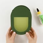Eine grüne Papierschablone wird von zwei Händen gehalten. Rechts liegt eine Klebstoffflasche mit gelbem Verschluss.