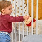 Ein Kleinkind in rotem Oberteil streichelt einen Teddybär im Gitterbett. Der Hintergrund zeigt gepunktete Stoffe.