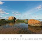 „Meeresstille“, Kalender mit großen runden Steinen im ruhigen Wasser, umgeben von Schilf, unter blauem Himmel mit Wolken.