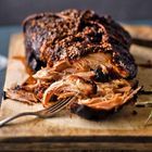 Saftiges, zerkleinertes Fleisch mit knuspriger Oberfläche auf einem Holzbrett, ein Metallgabel ist in das Fleisch gesteckt.