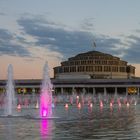 Text: „visit Wroclaw.eu“; Mehrstöckige Rotunde; davor ein Brunnen mit bunten Lichtern und Wasserspielen im Abendlicht.