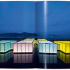 Leuchtende, farbige Kuben spiegeln sich im Wasser vor einem See mit Bergen im Hintergrund bei Dämmerung.