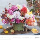 November. Ein prachtvoller Blumenstrauß in einem grünen Topf, umgeben von herbstlichem Laub und Früchten auf einem Tisch.