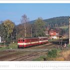 "NOVEMBER" steht unten. Ein roter Zug fährt durch eine ländliche Gegend mit Bäumen, Häusern und einem Motorradfahrer in der Nähe.