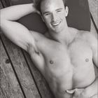 11. Ein Mann liegt lächelnd mit nacktem Oberkörper auf einem Holzboden, Arm hinter dem Kopf.