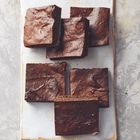 Sechs quadratische Brownies auf einer rechteckigen, weißen Platte. Der Hintergrund ist eine graue Steinoberfläche.