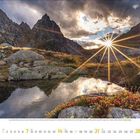 11 Nov | Ein Bergpanorama mit einem See im Vordergrund, Bergen im Hintergrund und strahlender Sonne hinter den Gipfeln.