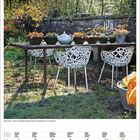 Kalender im November. Ein Tisch im Garten mit Dekorationen, umgeben von weißen Stühlen, Baum im Hintergrund.