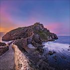 Eine gepflasterte Brücke führt zu einer felsigen Insel mit einer Kapelle unter einem farbenfrohen Sonnenuntergangshimmel.