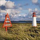 Ein rotes, dreieckiges maritimes Zeichen steht neben einem weißen Leuchtturm, umgeben von Dünen und Graslandschaft.